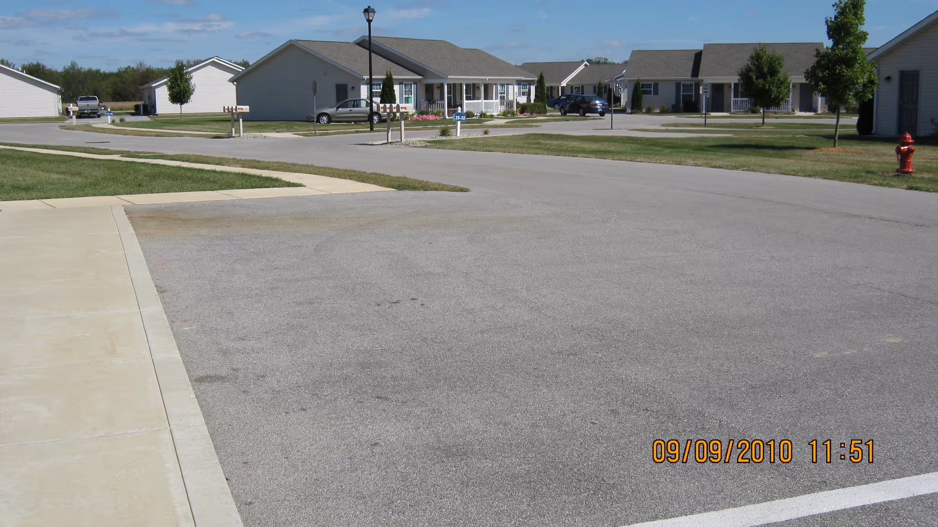 View of a quiet residential street in a senior living community with single-story houses, green lawns, a sidewalk, a fire hydrant, and a clear blue sky.