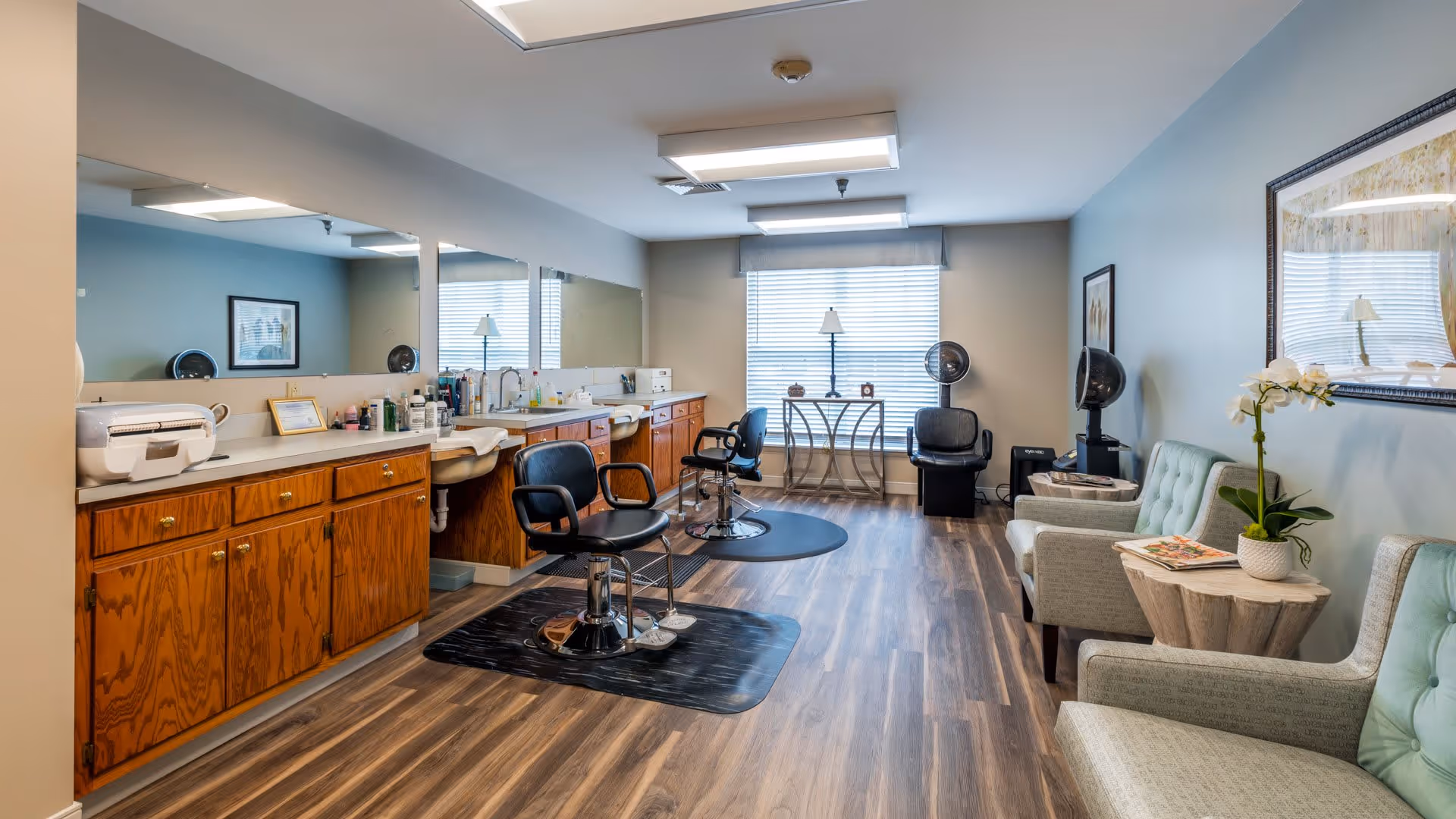 A well-lit salon area inside a senior living facility with wooden cabinets and countertops along the left wall, two black salon chairs on mats in front of large mirrors, and a seating area with three cushioned chairs and a small table with a potted plant on the right. A window with blinds and a small table with a lamp are visible at the far end of the room.