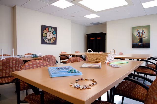 A bright communal dining area with long wooden tables, red patterned chairs, and wall art.