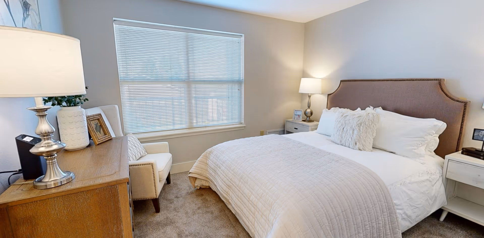 A cozy bedroom with a large bed featuring a beige upholstered headboard, white bedding, and a fluffy decorative pillow. There are two bedside tables with lamps on each side of the bed. A wooden dresser with a lamp, framed photo, and decorative items is positioned against the wall near a window with closed blinds. A white armchair is placed next to the dresser.