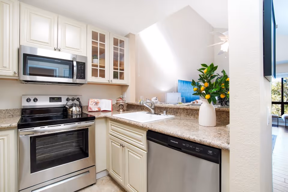 Modern kitchen with stainless steel appliances including a microwave, stove, and dishwasher. The kitchen features cream-colored cabinets, granite countertops, a white sink with a faucet, and a vase with green leaves and yellow fruit on the counter. In the background, part of a living area with a blue painting and large windows is visible.