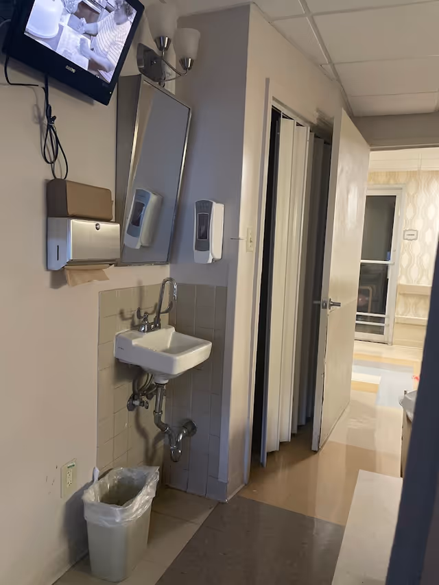 Small hand-washing station with a wall-mounted sink, mirror, soap and paper towel dispensers in a facility hallway.