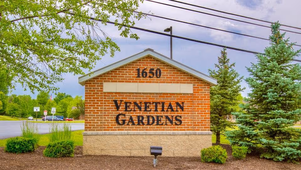 A brick sign with the address number 1650 and the name Venetian Gardens in black letters, surrounded by green shrubs and trees with a parking lot and street visible in the background under a partly cloudy sky.