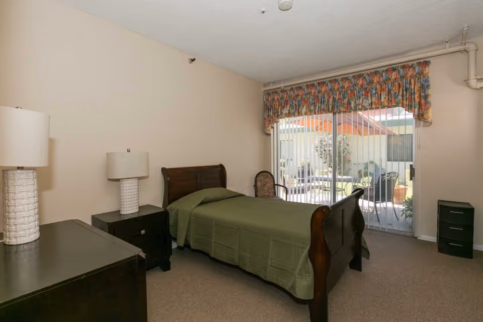 A bedroom with a single wooden bed covered with a green bedspread, two matching bedside tables with white lamps, a chair, and a large sliding glass door with vertical blinds and floral valance, leading to an outdoor patio area.
