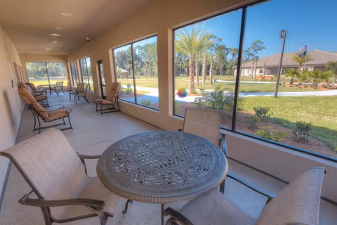 A covered outdoor patio area with several cushioned chairs and a round metal table. Large windows provide a view of a landscaped garden with palm trees and a building in the background under a clear blue sky.