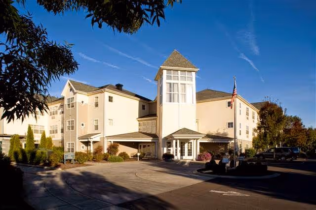 Exterior view of a multi-story senior living facility building with a prominent tower-like entrance, surrounded by landscaping and a driveway under a clear blue sky.