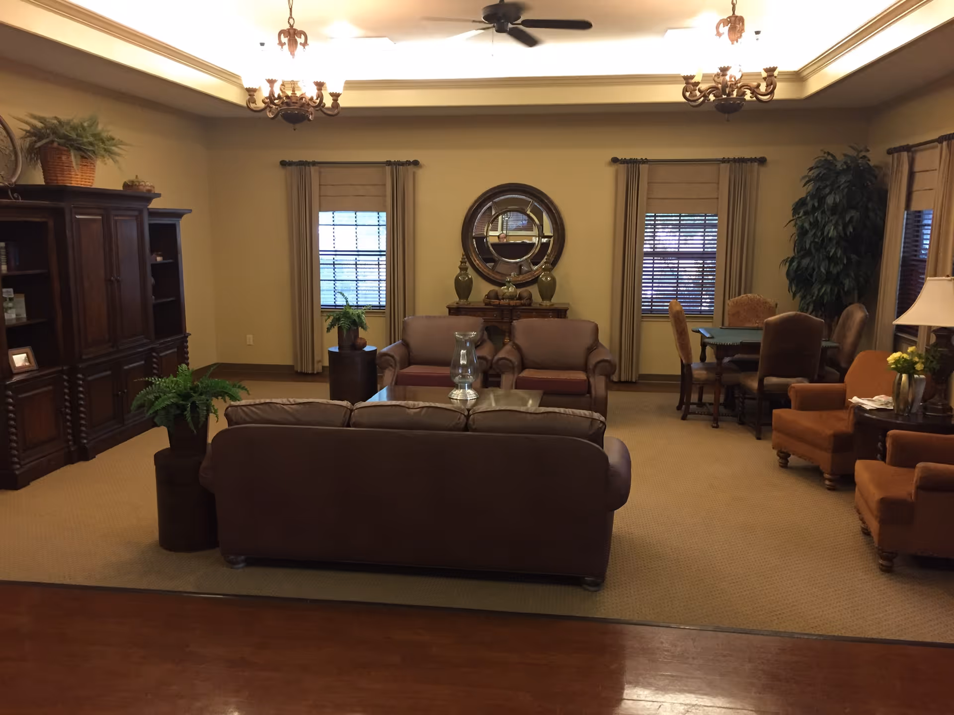 A cozy living room area with brown leather sofas and armchairs arranged around a glass coffee table. The room has beige walls, two windows with blinds and curtains, a large wooden cabinet on the left, and a round mirror above a small wooden table with decorative items. There are also plants and a ceiling fan with lights.