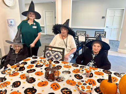 Three elderly women and a staff member dressed in witch hats sitting and standing around a table decorated with a Halloween-themed tablecloth featuring pumpkins, bats, and stars in a senior living facility dining or common area.