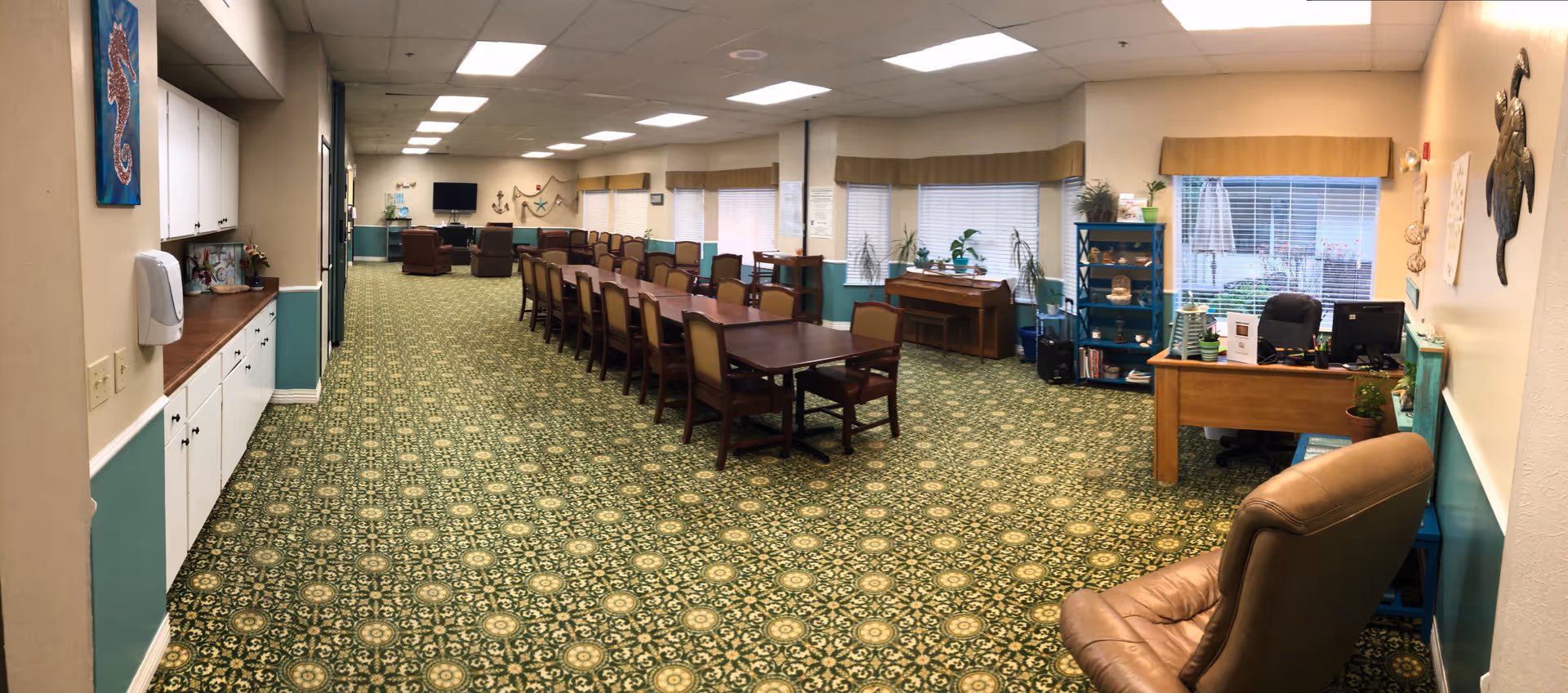 A long room with a patterned green carpet and beige walls, featuring a large wooden table with multiple chairs in the center. On the right side, there is a desk with a computer and office chair, a blue shelving unit with plants and decorative items, and a piano near the windows. The left side has white cabinets with a wooden countertop and a wall-mounted hand sanitizer dispenser. The room is well-lit with ceiling lights and has windows with blinds along the right wall.