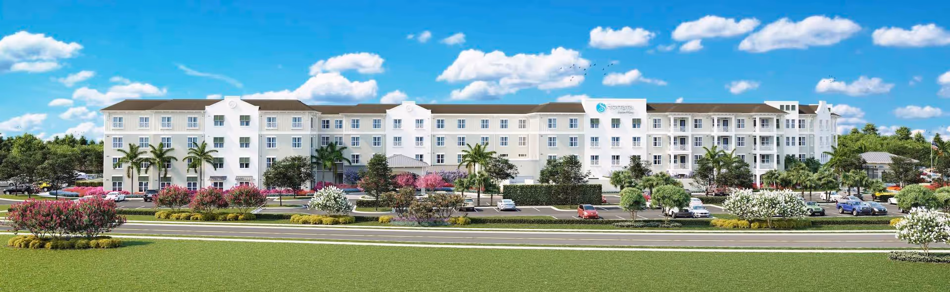 Long white four-story senior living building with palm trees, landscaped grounds and a parking area under a blue sky.