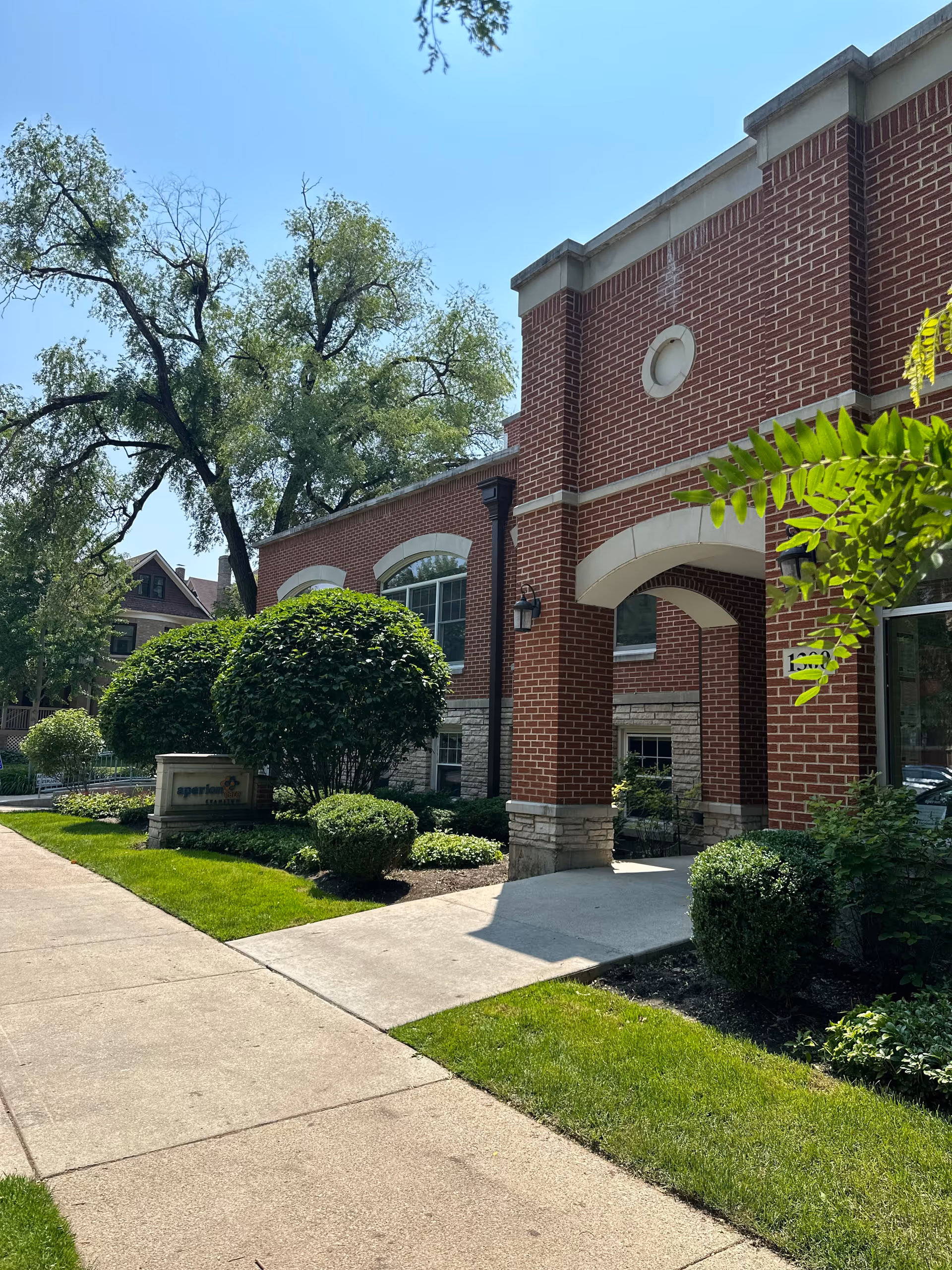 Exterior view of a red brick building with arched windows and an entrance archway, surrounded by neatly trimmed bushes and green grass under a clear blue sky.