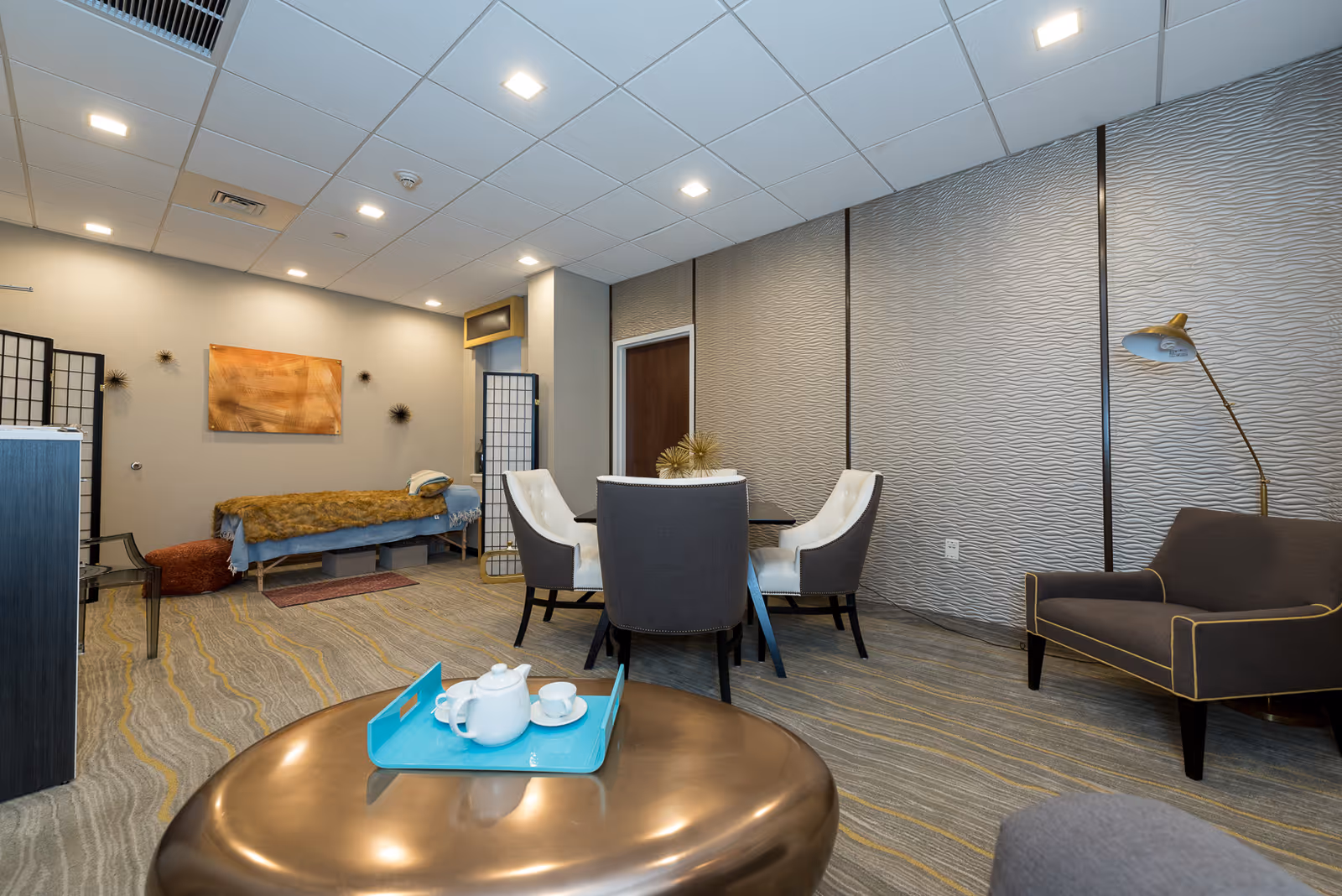 A modern interior room with a round table surrounded by four chairs, a comfortable armchair with yellow piping, a floor lamp, and a massage table covered with a brown blanket in the background. The room has textured walls, a patterned carpet, and ceiling lights.