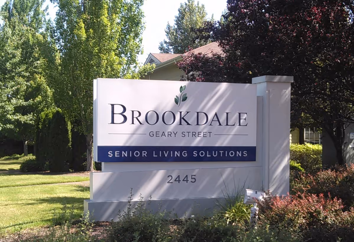 Outdoor view of a white sign for Brookdale Geary Street Senior Living Solutions with the address 2445, surrounded by green trees and bushes under a clear sky.