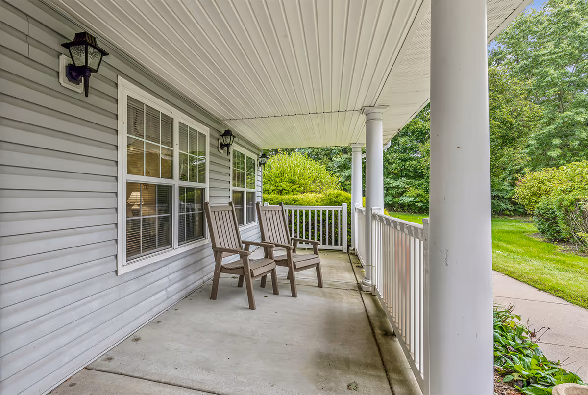 Covered porch area with two wooden chairs facing outward, white railing, and gray siding on the building. There are two windows with white frames and black outdoor wall lanterns mounted on the wall. Green bushes and trees are visible in the background.