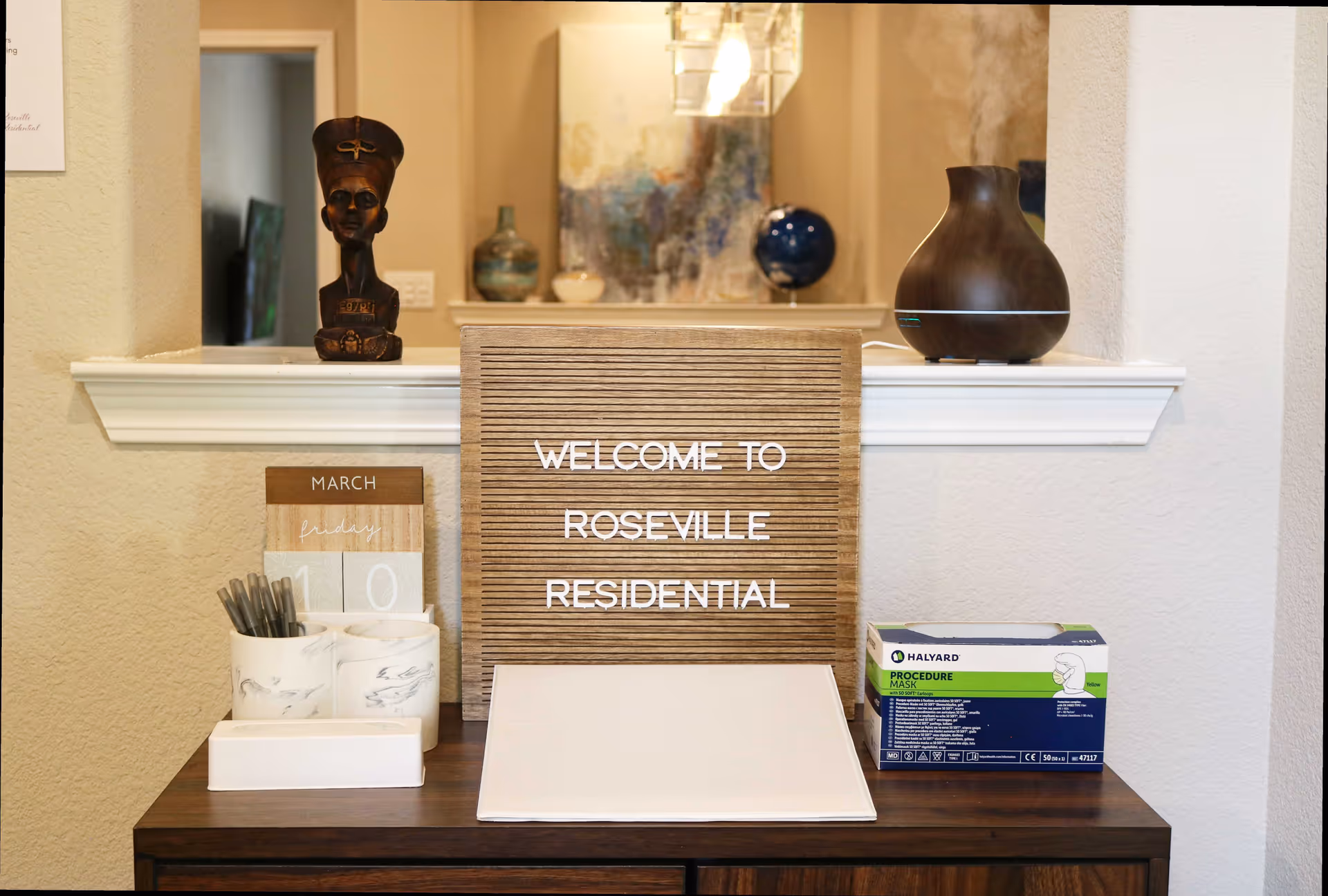 A reception table with a letterboard reading "WELCOME TO ROSEVILLE RESIDENTIAL" surrounded by decorative items, a small calendar, pens, and a box of procedure masks.