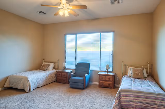 A bright bedroom with two single beds on opposite sides of the room, each with a nightstand and lamp. A blue recliner chair is positioned between the beds in front of a large window with blinds. The room has beige walls, carpeted floor, and a ceiling fan with lights.