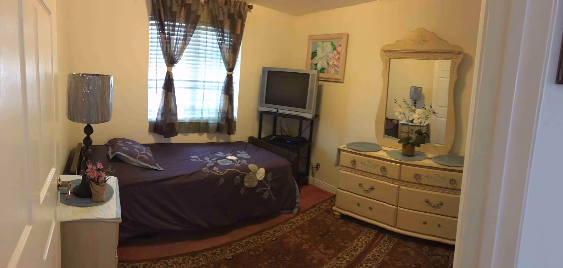 A small bedroom with a single bed covered in a dark floral bedspread, a nightstand with a lamp and a small potted plant, a window with sheer brown curtains, a TV on a stand, and a cream-colored dresser with a large mirror and decorative items on top.