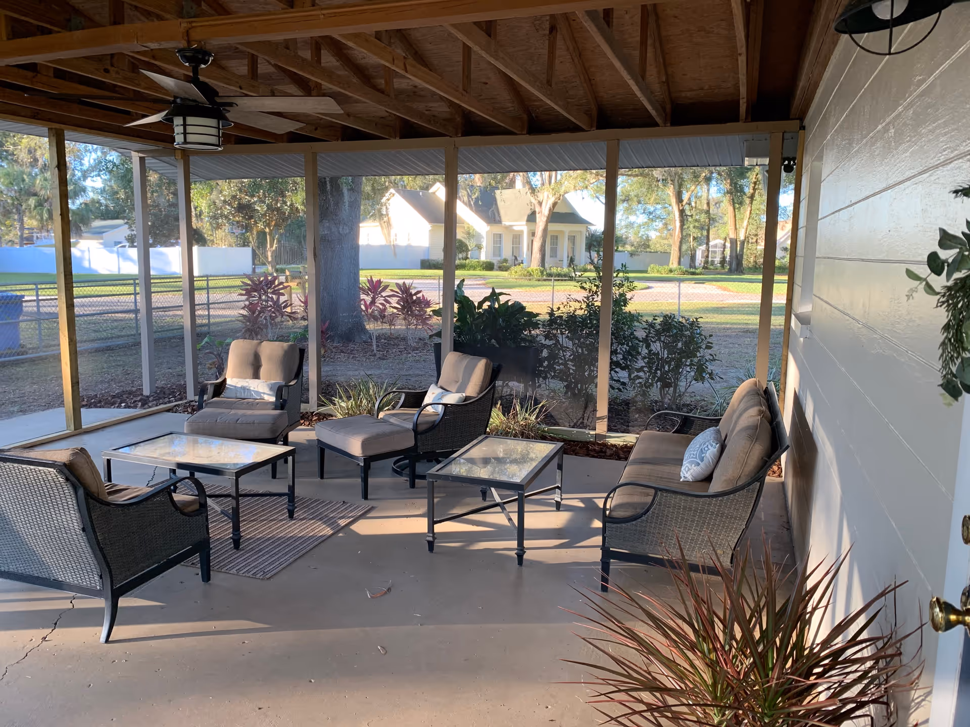 A covered outdoor patio area with cushioned wicker chairs, a loveseat, two glass-top tables, and a ceiling fan. The patio is screened in and overlooks a yard with trees, shrubs, and a white fence. A house is visible in the background.