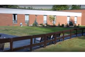 Exterior view of a single-story brick nursing facility with a grassy lawn, a walkway ramp with wooden railing and benches.