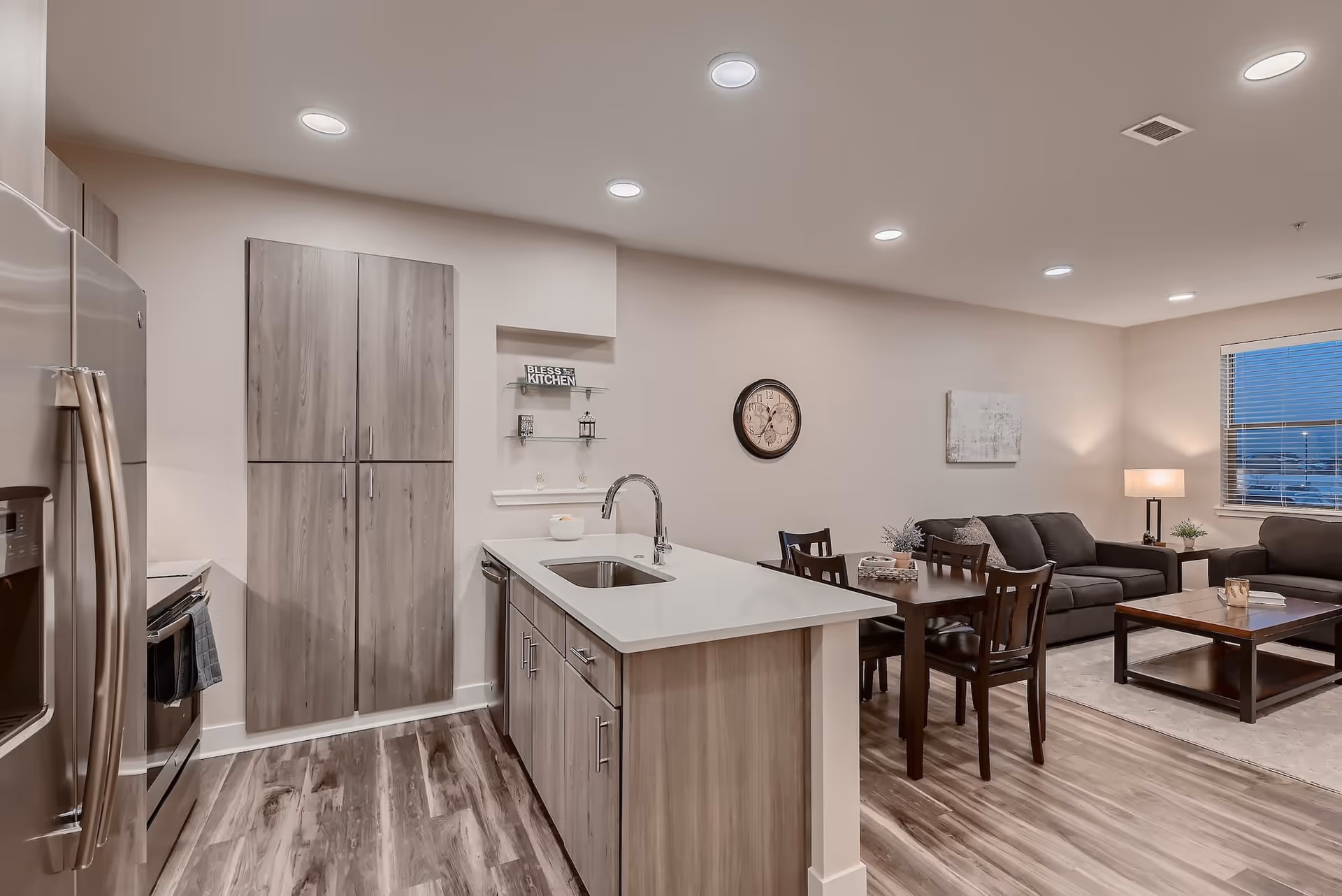 Modern open-concept kitchen and living room area with wood flooring. The kitchen features stainless steel appliances, a large island with a sink, and wood cabinets. The living room has dark sofas, a coffee table, a side table with a lamp, and a window with blinds. A dining table with chairs is positioned between the kitchen and living room. A wall clock and decorative items are visible on the walls and shelves.