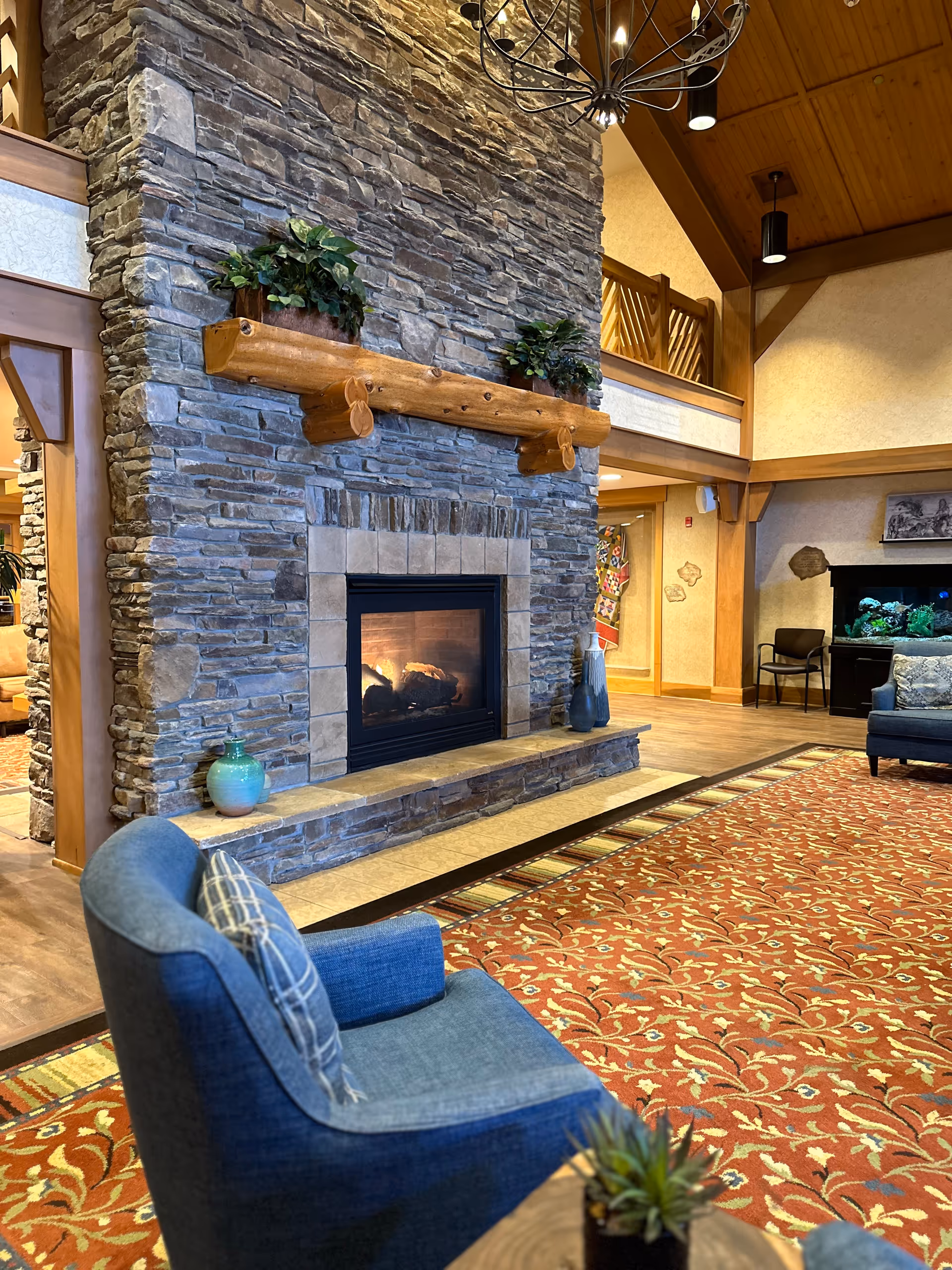 Cozy senior living facility common area with a large stone fireplace featuring a wooden mantel decorated with two potted plants. In front of the fireplace is a blue upholstered chair with a plaid pillow. The room has a high wooden ceiling with exposed beams and a chandelier. A patterned red and orange carpet covers the floor, and there is a fish tank and additional seating visible in the background.