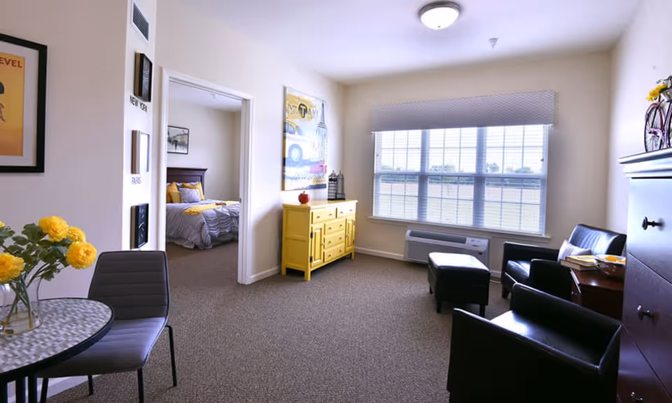 A bright senior living room with a large window covered by a valance, a yellow sideboard against the wall, two black leather chairs with a matching ottoman, and a small round table with two chairs and a vase of yellow flowers. A doorway leads to a bedroom with a bed and yellow pillows.