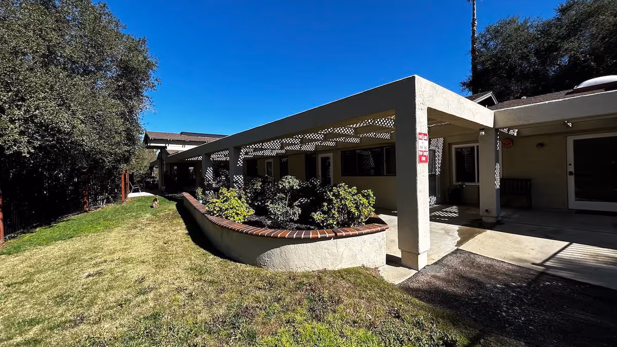 Outdoor view of a senior living facility showing a covered patio area with a raised garden bed filled with plants. The building has beige walls and several windows, with a grassy lawn and trees on the left side under a clear blue sky.