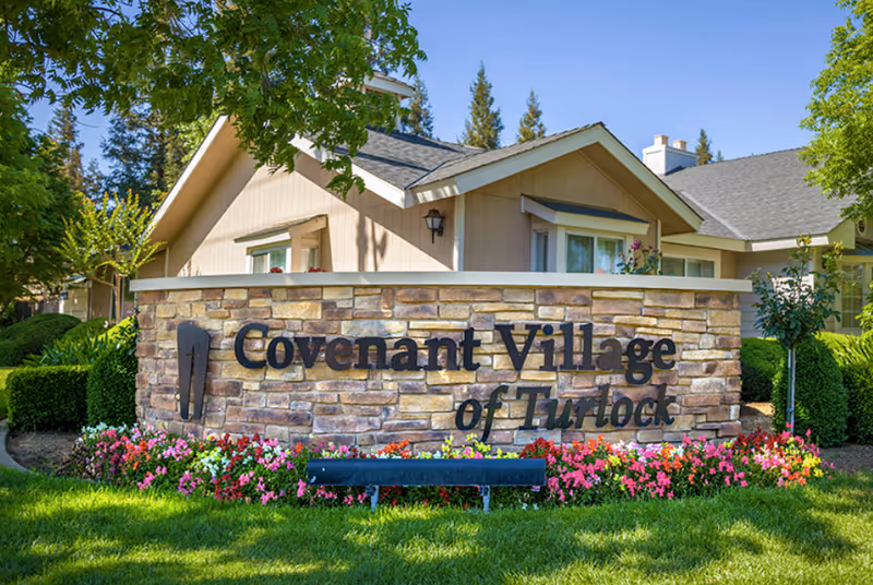 Stone sign with the text 'Covenant Village of Turlock' surrounded by colorful flowers and green grass, with a beige residential building and trees in the background under a clear blue sky.