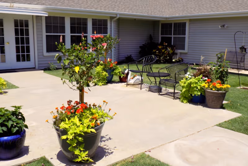 Outdoor patio area at Mackenzie Court featuring potted plants with colorful flowers, metal chairs and a bench on a concrete surface, adjacent to a building with windows and a door.