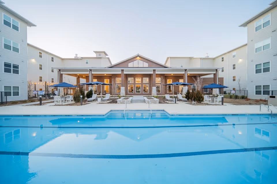 Outdoor swimming pool in front of a multi-story residential building with a covered patio area featuring tables, chairs, and blue umbrellas at Sage Highland Creek facility.