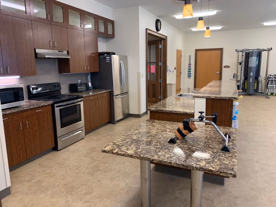 Interior view of a kitchen area with wooden cabinets, a stainless steel refrigerator, stove, microwave, and granite countertops. There is a long counter with a sink and a hand exercise device on the countertop. In the background, there is exercise equipment and a beige tiled floor.