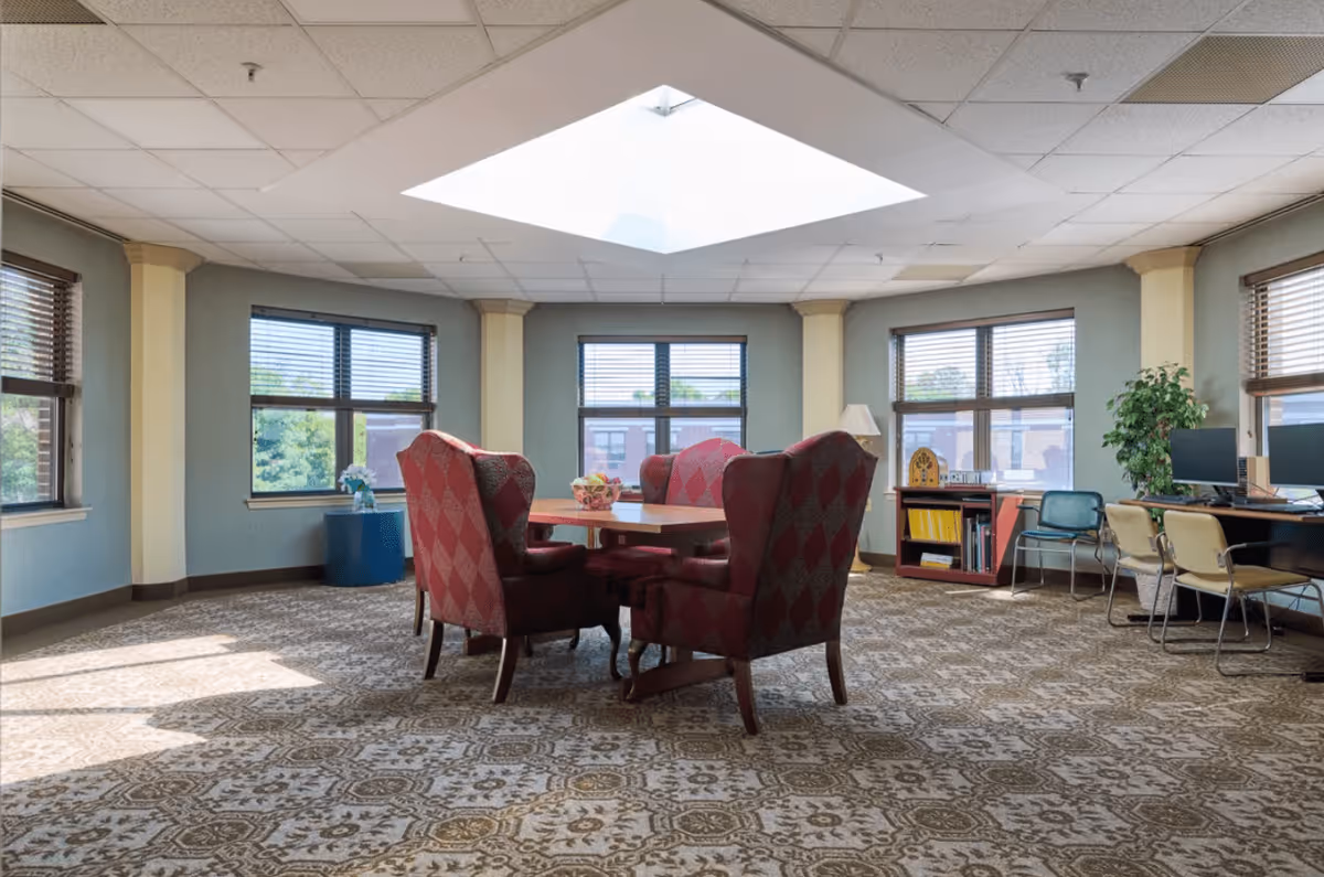 A bright room with large windows on three sides, featuring a skylight in the ceiling. In the center, there is a wooden table surrounded by four red patterned upholstered armchairs. To the right, there is a desk with two computer monitors and several chairs. The room has a patterned carpet and light-colored walls with columns between the windows.