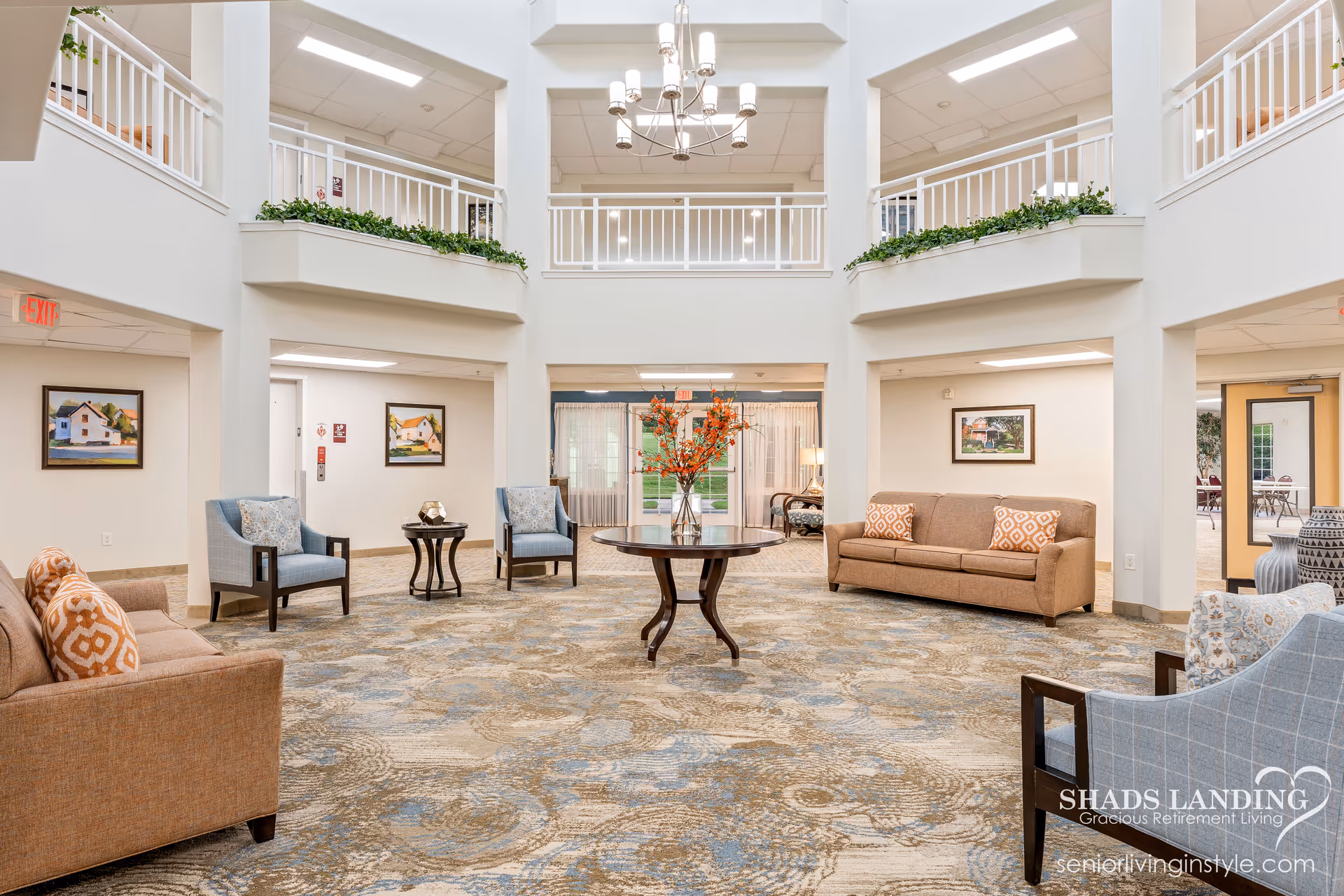 A spacious and well-lit retirement living common area with beige and blue patterned carpet, two brown sofas with orange patterned pillows, two blue armchairs, a round wooden table with a vase of orange flowers in the center, white railings on the upper level, and framed paintings on the walls.