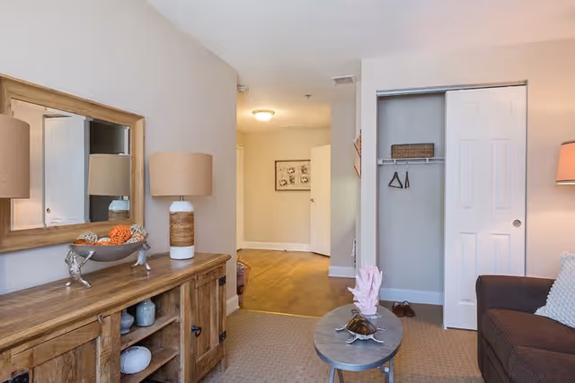 A cozy living room area featuring a wooden sideboard with a lamp and decorative items, a small oval coffee table with seashell decorations, a brown sofa with a white pillow, and an open closet with hangers and a basket. The room has beige walls and carpeted flooring, with a view into a hallway with wooden flooring and a framed picture on the wall.