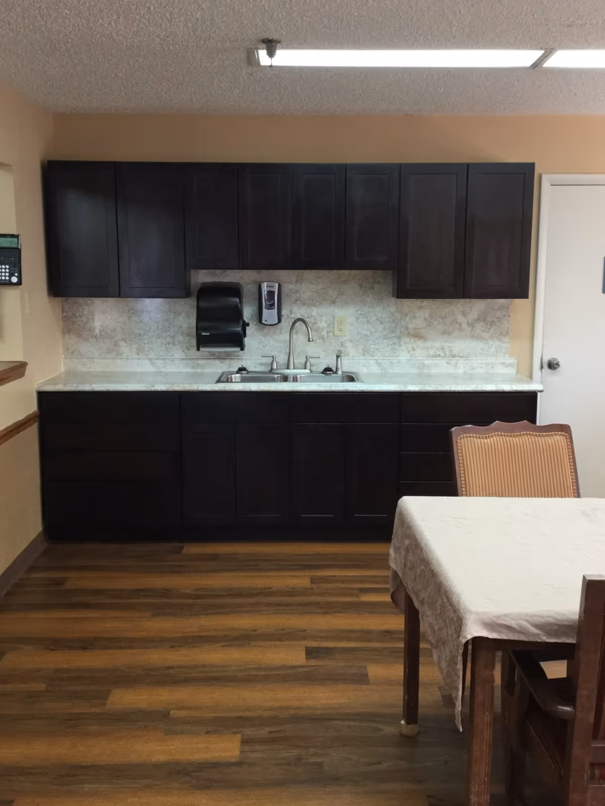 Small kitchenette with dark upper and lower cabinets, a double sink, and a table with chairs on wood flooring.