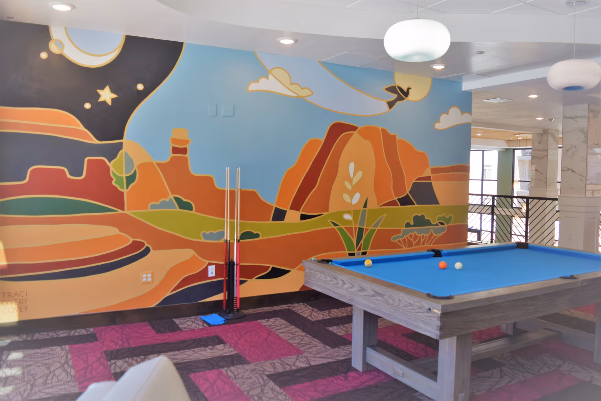 A recreational room with a pool table featuring a blue felt surface and several billiard balls on it. The room has a colorful mural on the wall depicting a stylized desert landscape with rock formations, plants, and a night sky with stars and a moon. There are pool cues standing upright against the wall near the mural. The floor has a patterned carpet in shades of purple and gray, and the ceiling has recessed lighting and hanging round light fixtures.
