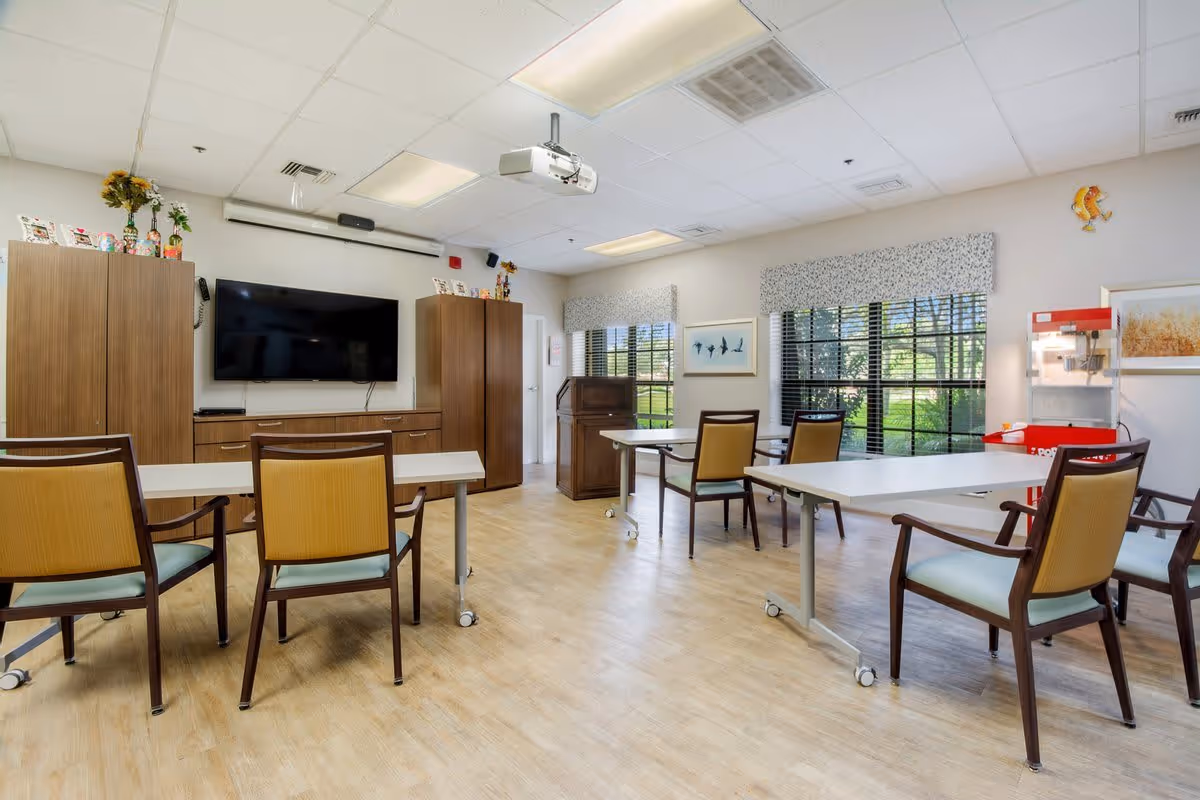 A bright and spacious common room with light wood flooring, several tables with chairs, a large flat-screen TV mounted on the wall, wooden cabinets, a popcorn machine, and large windows with patterned valances letting in natural light.