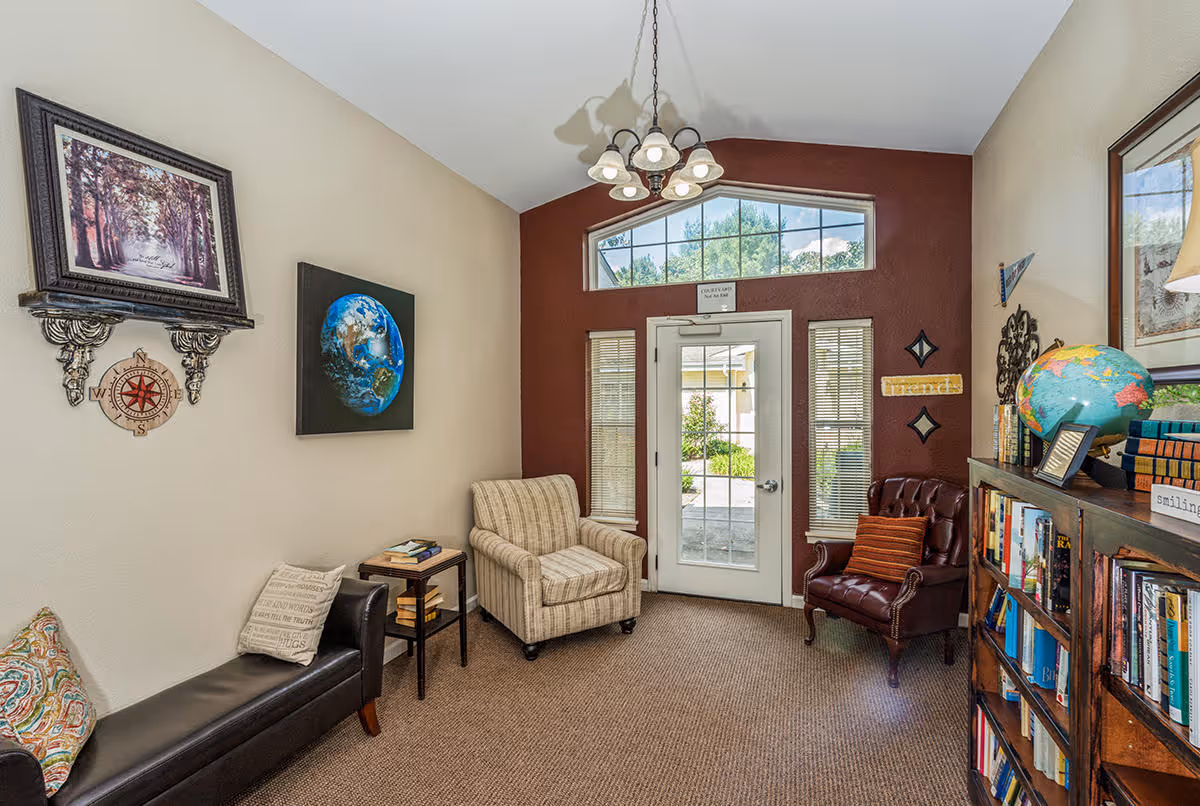 A cozy sitting area in Cedar Creek of Bedford featuring a beige armchair, a dark brown leather chair with an orange pillow, a black leather bench with decorative pillows, a small side table with books, and a wooden bookshelf filled with books and a globe. The room has beige and maroon walls, a glass door with sidelights and a transom window letting in natural light, and various wall decorations including framed pictures and a compass wall hanging.