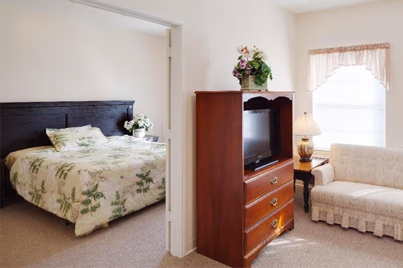 Bright suite interior with a bedroom visible through a doorway and a living area featuring a wooden TV cabinet, sofa, and side table with lamp.