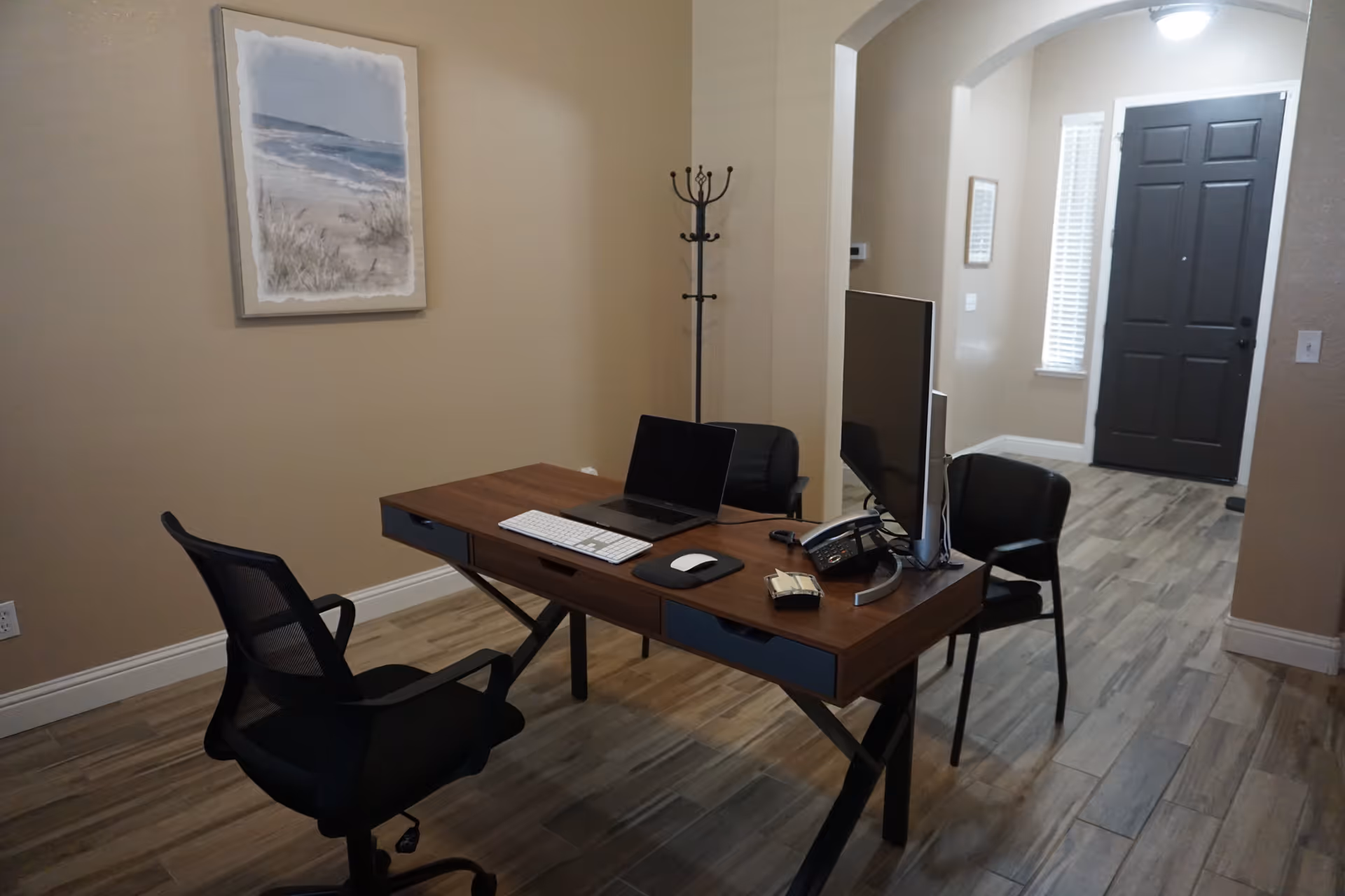 An office area with a wooden desk holding a laptop, keyboard, mouse, phone, and a large monitor. Two black chairs are positioned on either side of the desk. The room has beige walls, a coat rack, a framed beach painting, and a dark front door visible in the background.