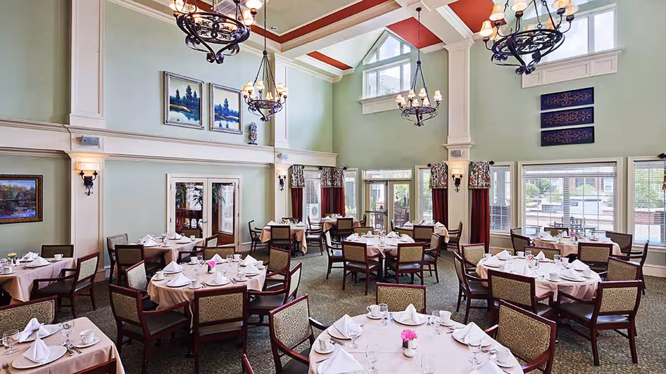 A spacious dining room with multiple round tables covered in light pink tablecloths, each set with white napkins, cups, glasses, and small flower vases. The room features high ceilings with decorative chandeliers, large windows with curtains, and light green walls adorned with framed artwork.