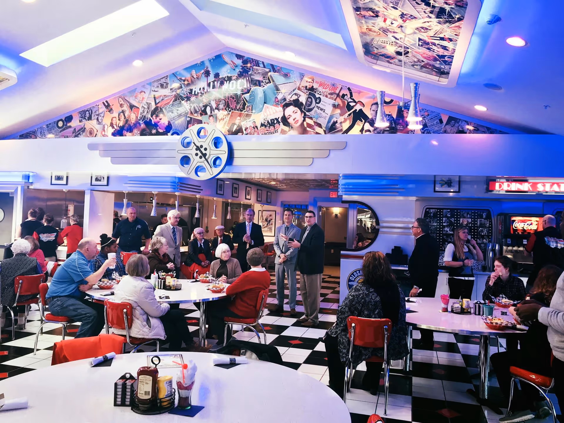 A retro-style diner interior with black and white checkered floor and red chairs. Several elderly people and a few younger adults are seated at round tables, eating and drinking. The walls and ceiling feature vintage movie and Hollywood-themed decorations, including a large film reel emblem. There is a drink station with a Coca-Cola dispenser on the right side.