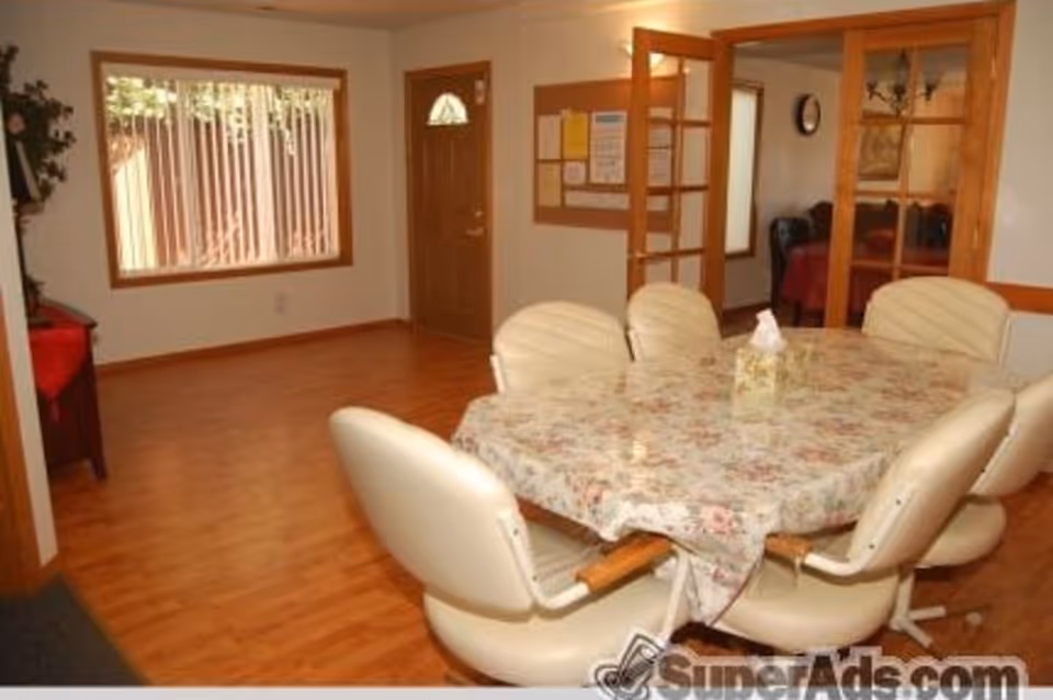 Interior dining room with a floral-covered table surrounded by beige swivel chairs on wood floors, a window and wooden door in the background.