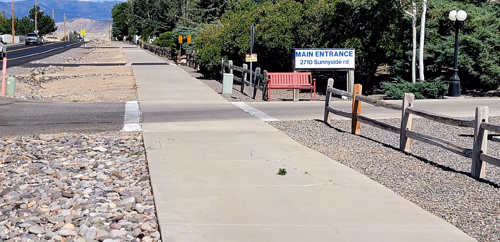 A concrete sidewalk running alongside a road with a rocky landscape on one side and a fenced gravel area with trees and bushes on the other. A red bench is positioned near a sign that reads 'MAIN ENTRANCE 2710 Sunnyside rd'. Mountains are visible in the background under a clear sky.