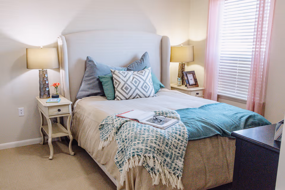 Well-lit bedroom with a neatly made bed, decorative pillows, bedside tables with lamps, and a window with pink curtains.