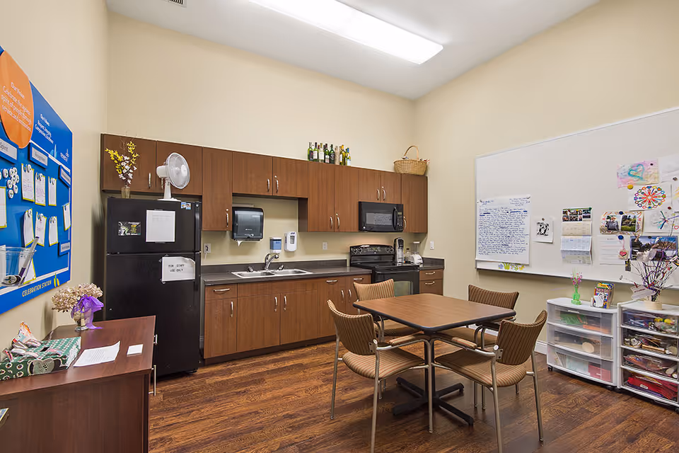 A small kitchen area with wooden cabinets, a black refrigerator, stove, microwave, and sink. There is a table with four chairs in the center of the room. On the walls, there is a large whiteboard with various papers and drawings pinned to it and a blue bulletin board with notes. The floor is wooden, and the room is well-lit with a ceiling light.