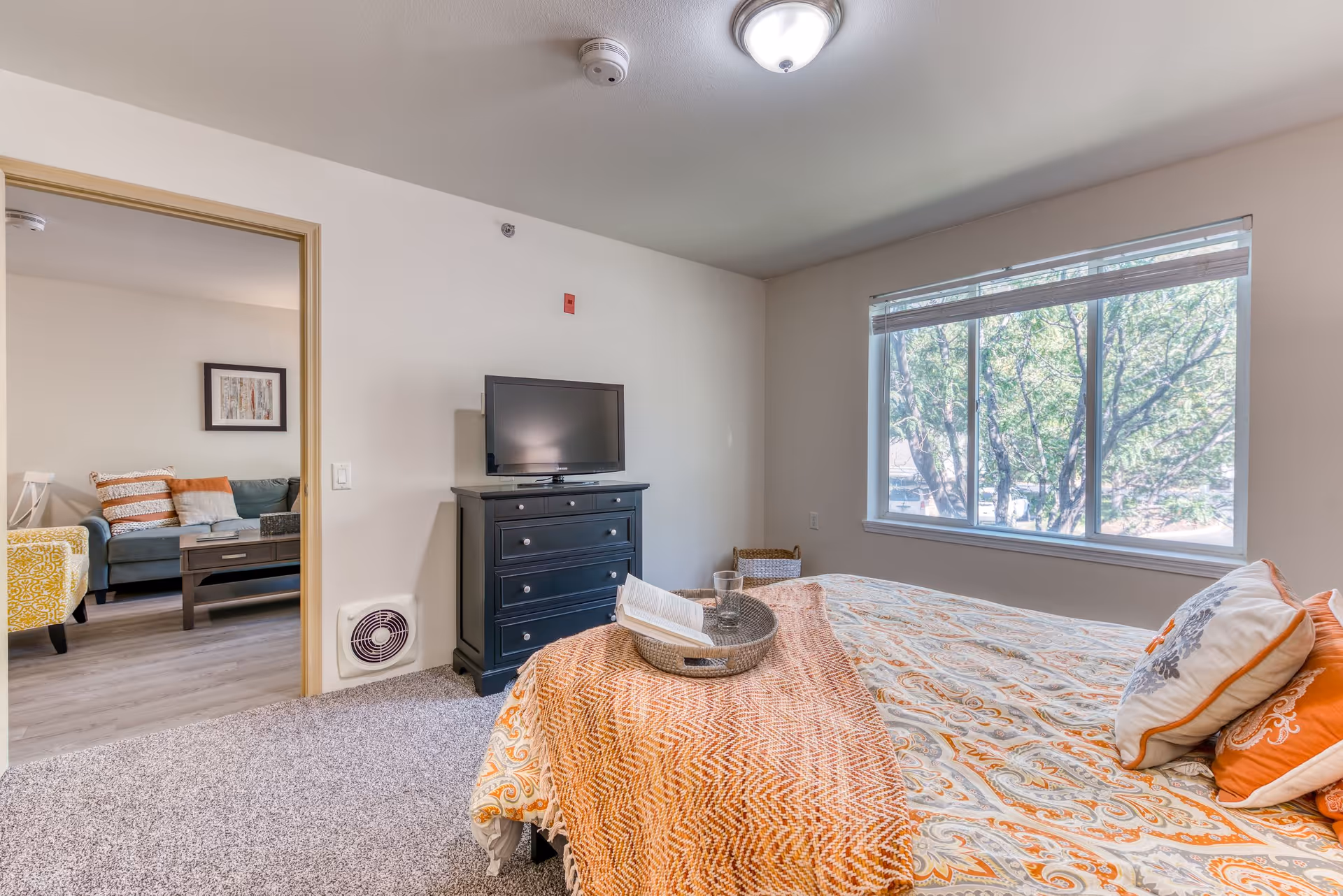 A bright bedroom with a large window showing trees outside. The bed is covered with a patterned bedspread and orange throw blanket, with several pillows. A black dresser with a flat-screen TV on top is against the wall. Through an open doorway, a living room with a sofa, coffee table, and armchair is visible.