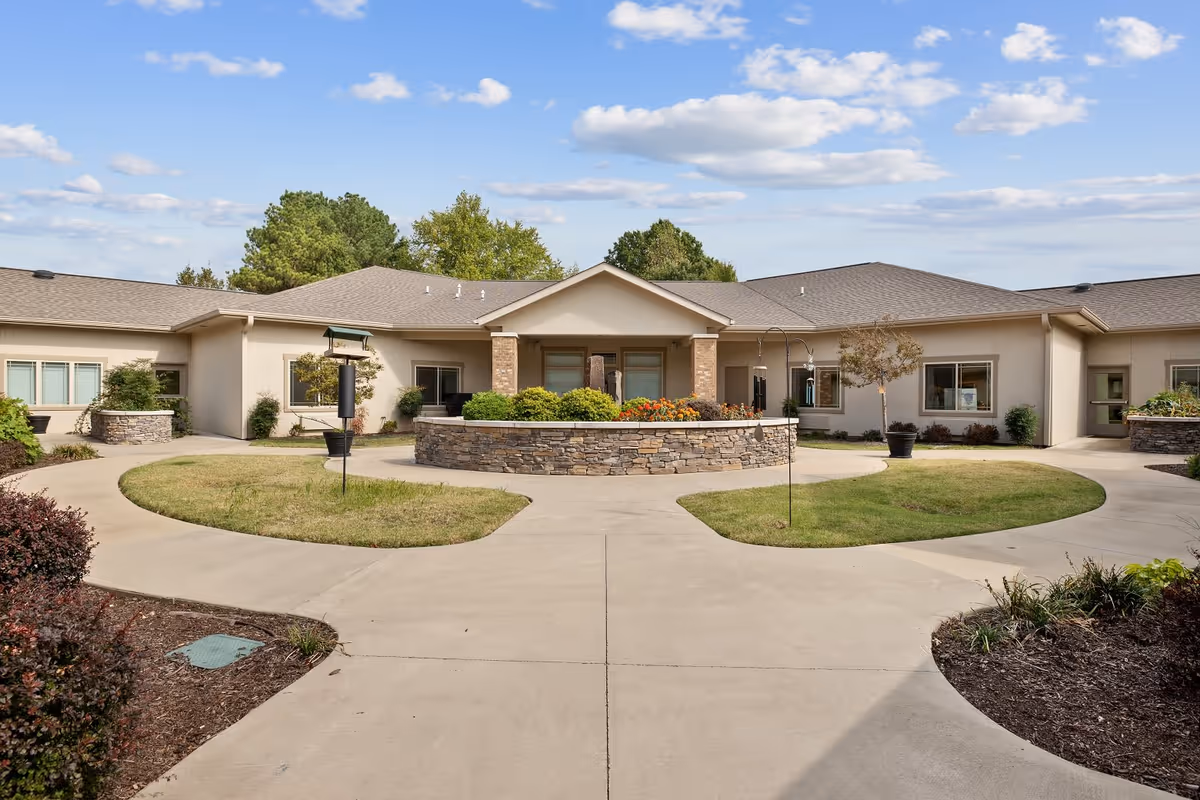 Exterior view of Magnolia Place Transitional Assisted Living and Memory Care building with a circular driveway, landscaped garden beds, and a partly cloudy sky.