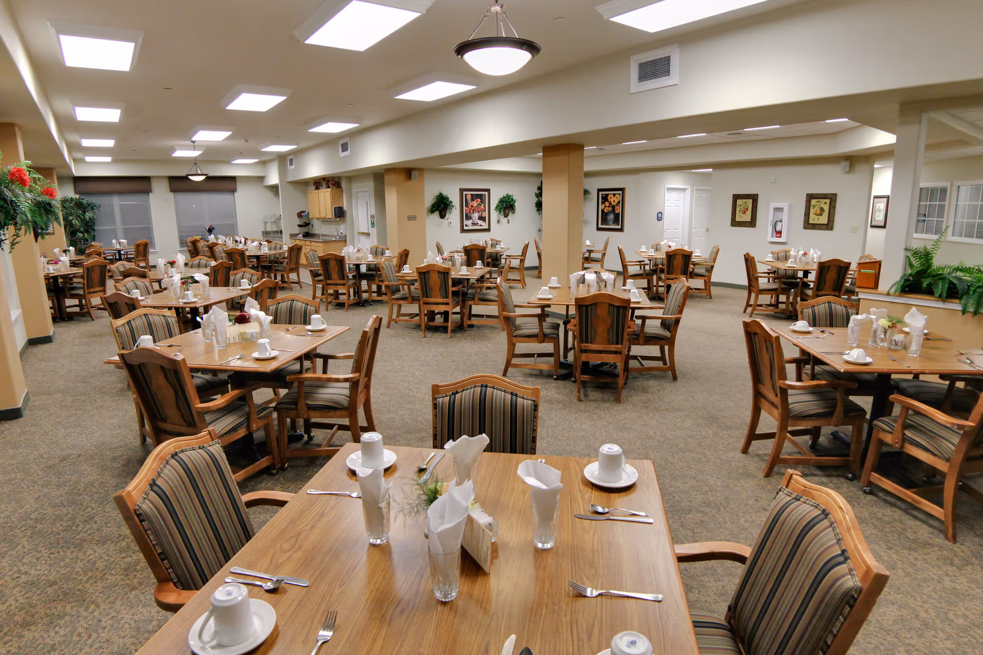 A spacious dining room with multiple wooden tables and chairs arranged neatly. Each table is set with white cups, saucers, utensils, and napkins. The room is well-lit with ceiling lights and decorated with framed pictures and green plants along the walls.