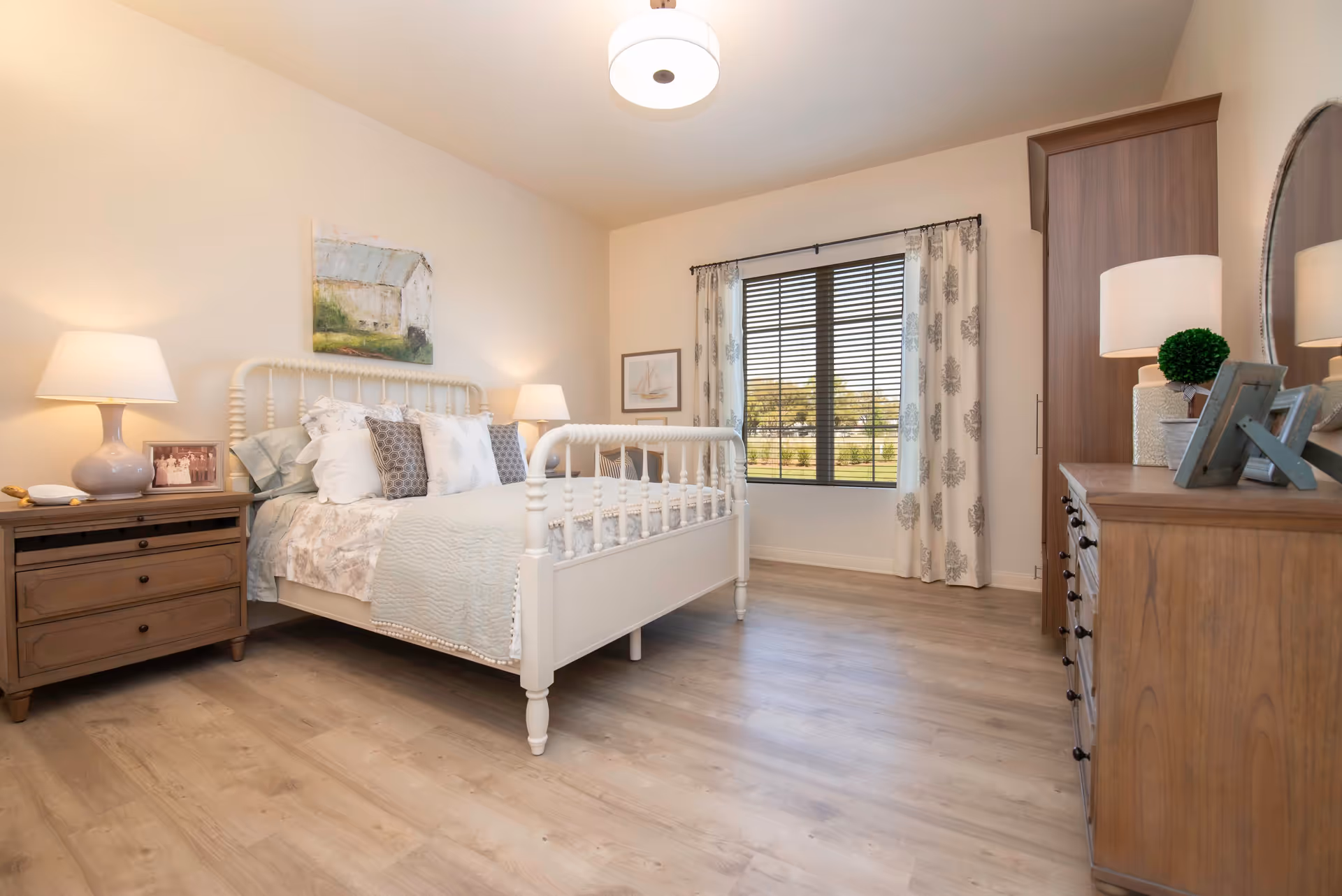 A bright and cozy bedroom with a white wooden bed frame, multiple pillows, and a light quilt. There are two bedside tables with lamps, a wooden dresser with a lamp and decorative items, a large window with patterned curtains and blinds, and light wood flooring.